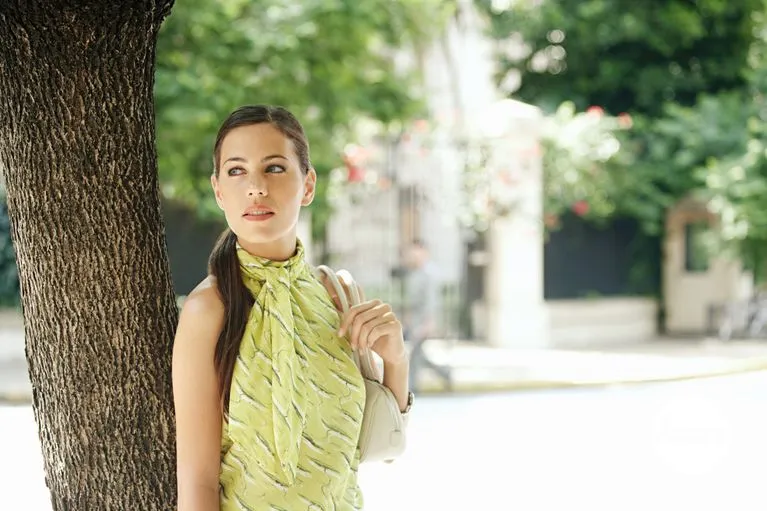 Attractive young businesswoman standing under a tree in the city, holding her handbag.
