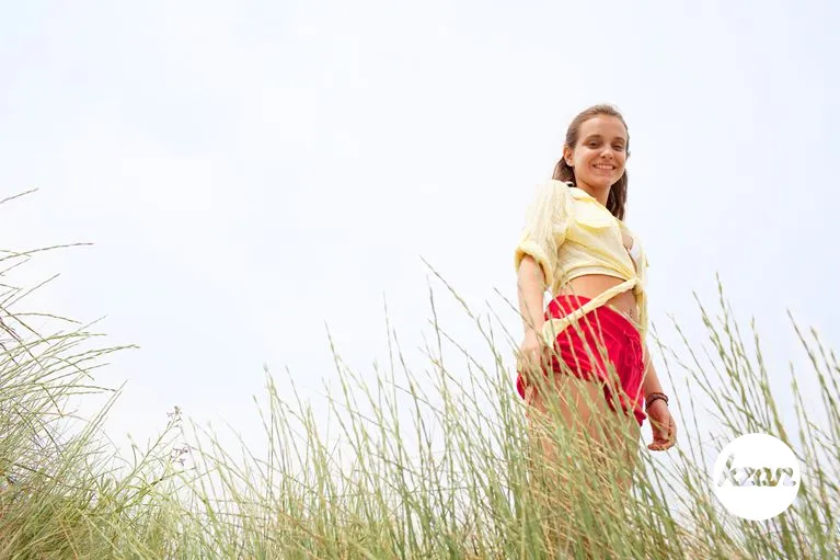 Beautiful healthy teenager tourist girl looking smiling at camera, on beach sand dunes with long green grass and sunny sky, enjoying summer holiday vacation, outdoors. Adolescent recreation lifestyle.