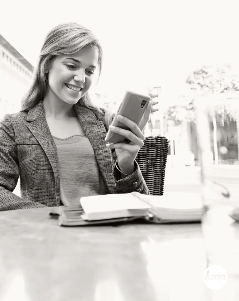 Black and white portrait of a beautiful business woman at a coffee shop terrace table holding a smart phone to work, smiling outdoors. Professional woman in city using technology. Smart girl lifestyle, sunny exterior.