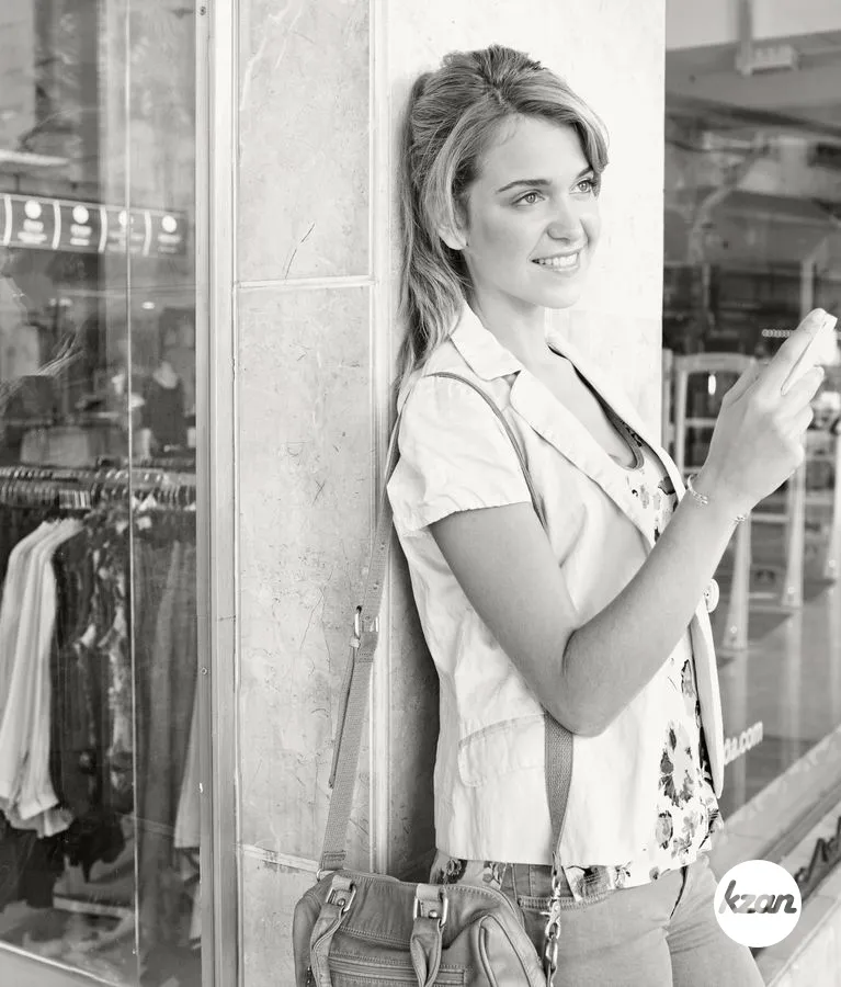 Black and white portrait of a beautiful young woman in a city shopping street, smiling using a smart phone. Tourist woman using technology, travel lifestyle. Shopping mall exterior with fashion store.