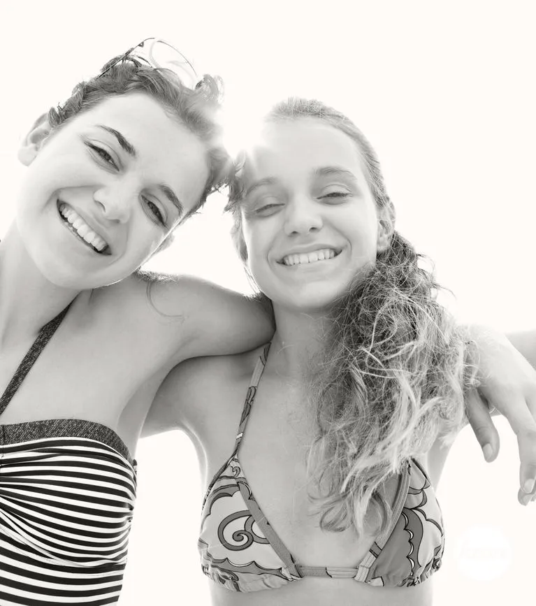 Black and white portrait of beautiful sisters on summer beach holiday joyful smiling at camera with heads together and expressions with sun rays filtering, outdoors. Travel fun recreational lifestyle.
