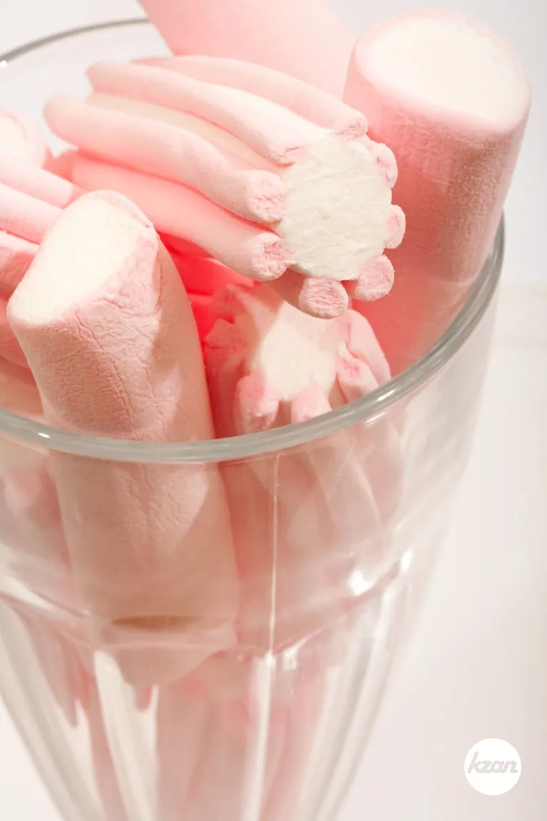 Close up detail still life view of a large glass full of candy sweet pink marshmallows isolated against a white background, interior. Comforting and naughty indulgence foods with sugar and fat. Sweet tooth party eating.