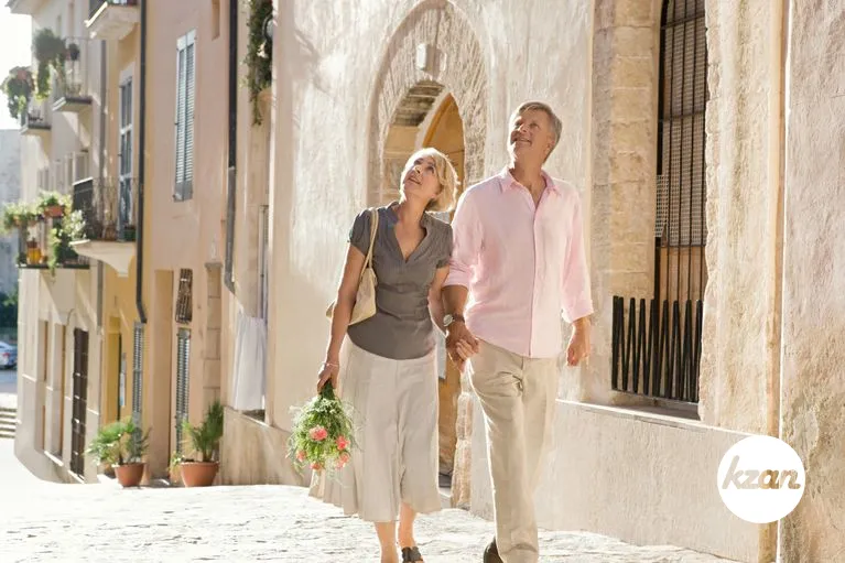 Mature tourist couple walking together holding hands and flower bouquet, visiting old city street architecture, outdoors. Senior mand and woman on holiday, leisure recreation lifestyle.