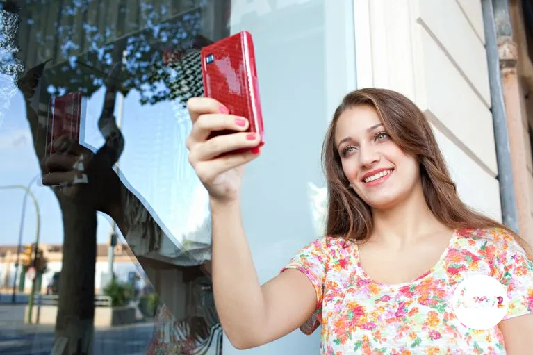 Portrait of a beautiful joyful young woman holding her hand up and using a smartphone mobile taking a selfie photo of herself during a sunny day, smiling. Lifestyle and technology outdoors.