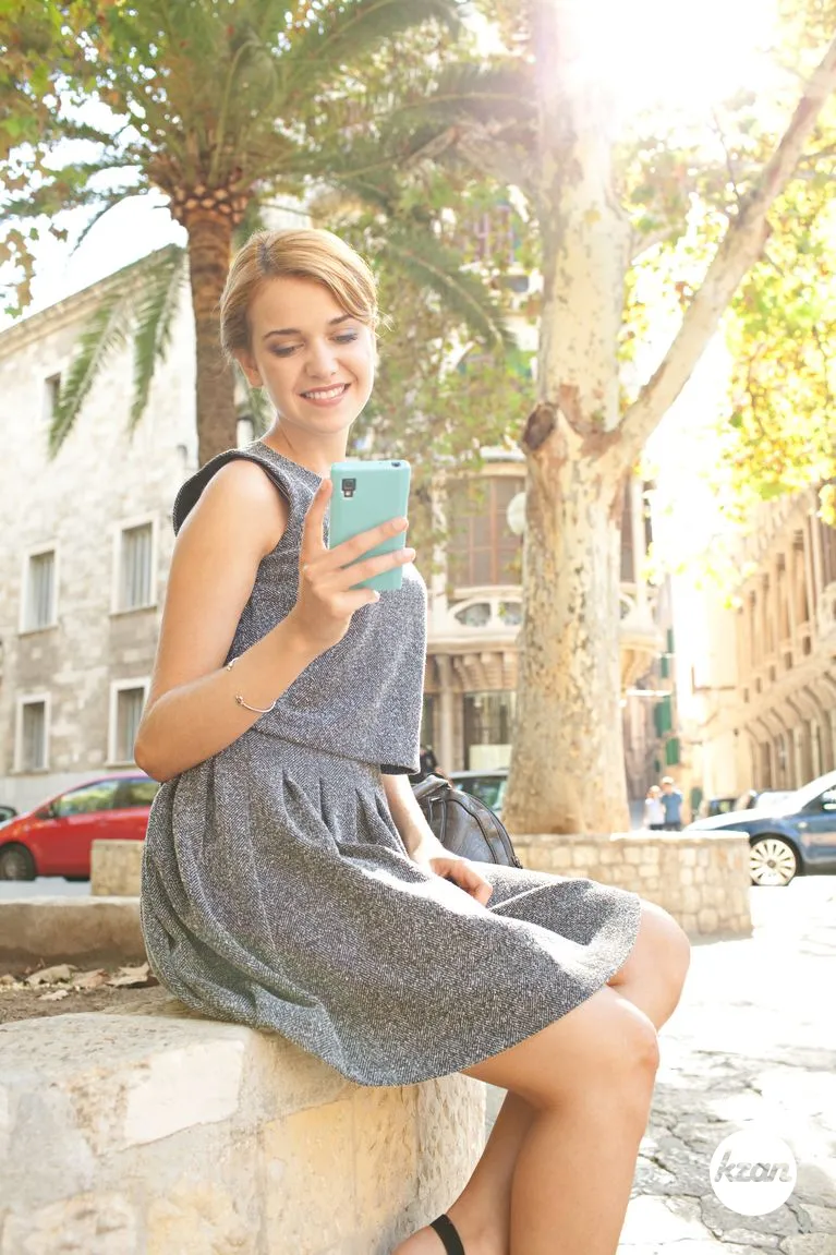 Portrait of beautiful professional young woman using a smart phone working in city with classic buildings outdoors, sunny day. Confident aspirational business woman sitting and smiling with technology, city lifestyle.