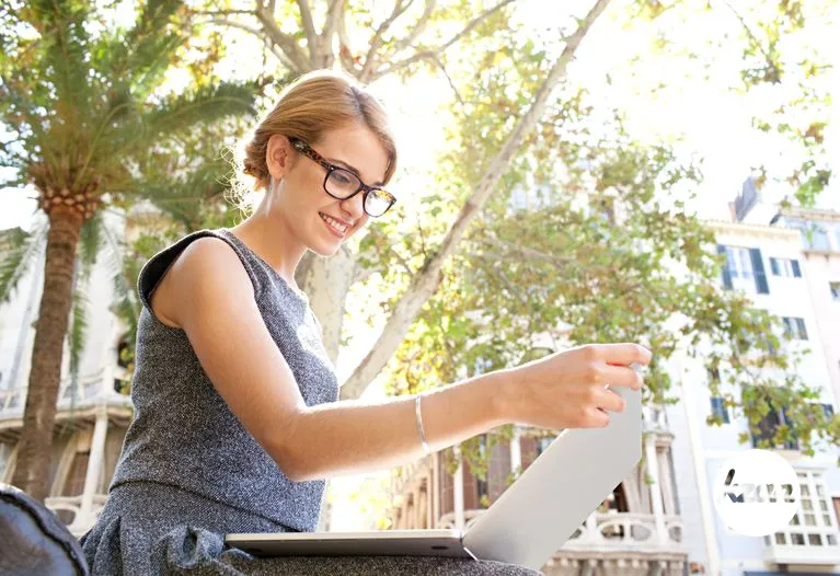 Side portrait of a young smart business woman using reading spectacles with laptop computer sitting in a city on a sunny day, smiling outdoors. Technology work and lifestyle, girl using glasses, exterior.