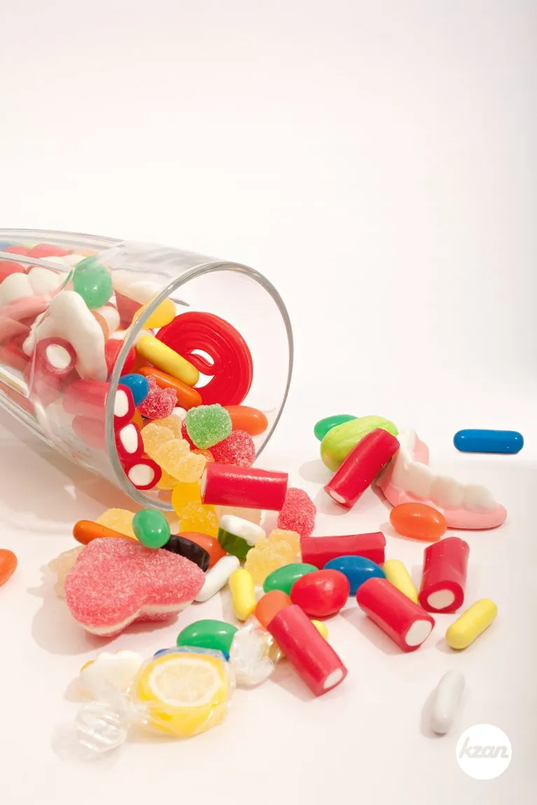 Still life of a glass container with different colorful candy sweets, fruity sticks, liquorice and jelly beans spilling out, isolated against a white background. Indulgence and naughty sugar and fat full food temptation, interior.