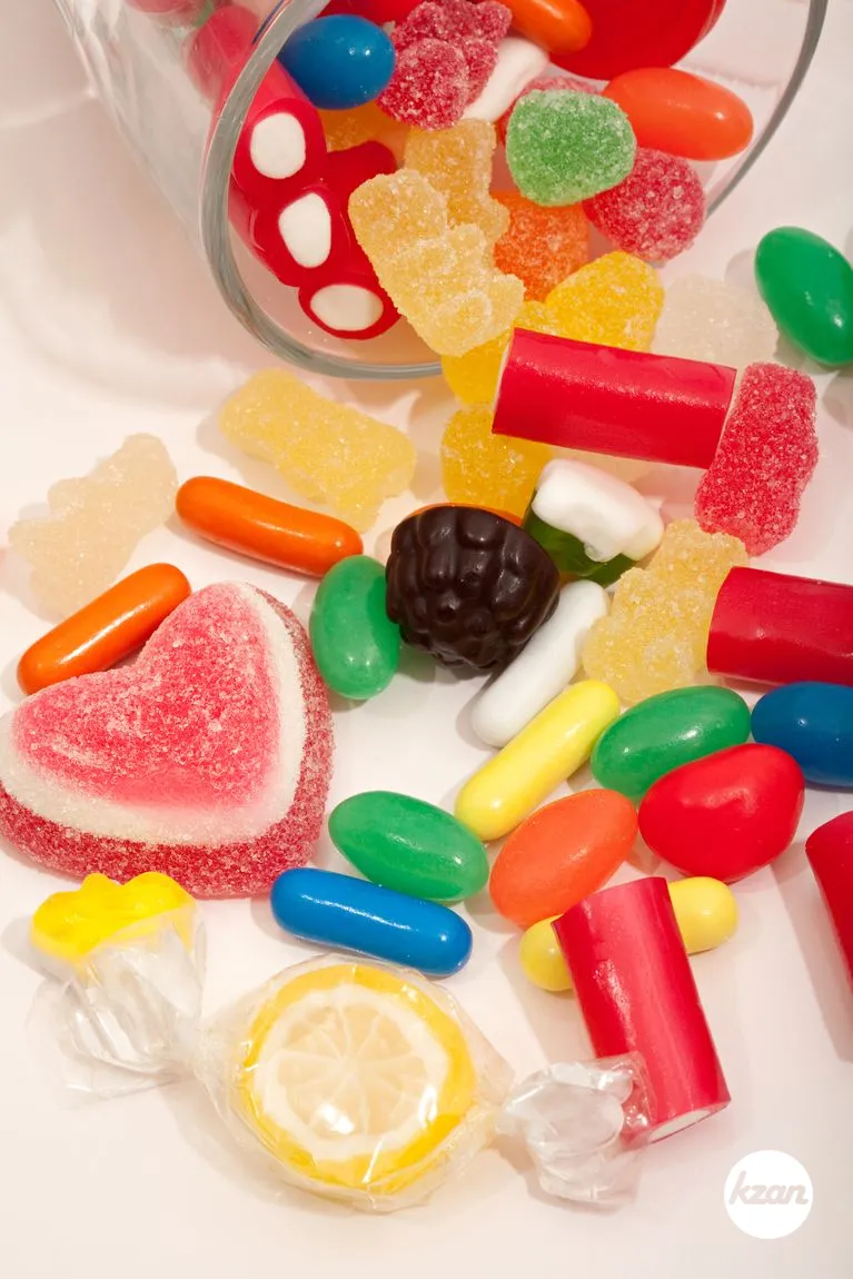 Still life of a glass container with different colorful candy sweets, liquorice and jelly beans spilling out isolated against a white background. Indulgence and naughty sugar and fat full food temptation, interior.