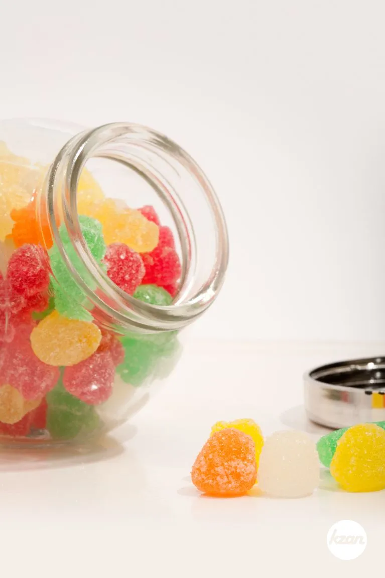 Still life of an open glass jar container with different colorful candy sweets and jelly beans isolated spilling out against a white background. Indulgence and naughty sugar and fat full food temptation.