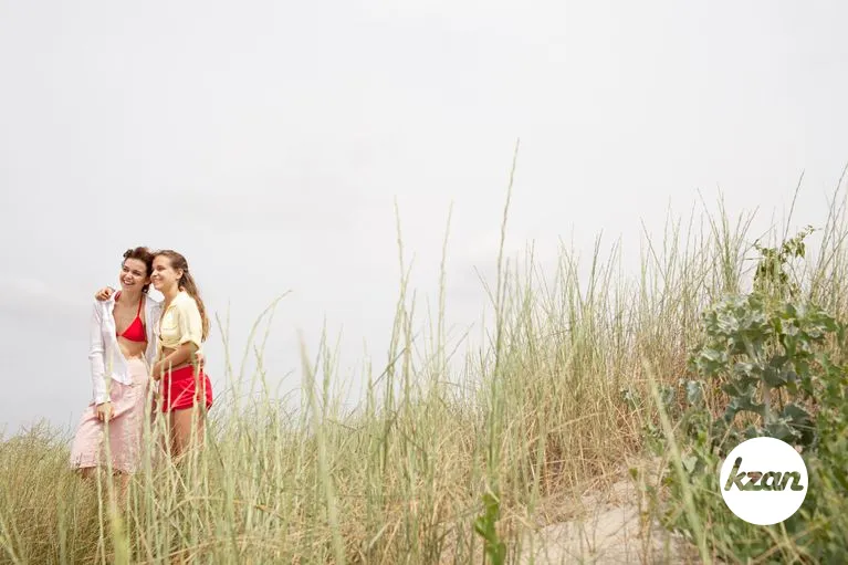 Two healthy girls friends relaxing together on beach dune's grass, smiling on summer holiday vacation, contemplating the sea, nature outdoors. Close females, healthy recreation lifestyle, coastal exterior.